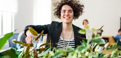 Eine Frau steht in einem Büro, Frau Grün, Pflanzen und gießt mit ihrem Glas Wasser die Blumen